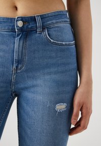 Μπλε ψηλόμεσα τζιν με φθαρμένο γόνατο, διαθέτοντας ασημένιο κουμπί, κλασικό σχέδιο πέντε τσεπών και ξεθωριασμένη υφή denim.