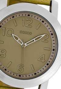 Montre ronde avec un cadran beige, boîtier en acier inoxydable argenté, grands chiffres et marqueurs noirs contrastants. Bracelet en cuir jaune.