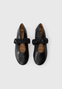 Een paar zwarte platte schoenen met opgeplooide elastische bandjes over de bovenkant, tan gekussende binnenzolen en ronde neuzen op een witte achtergrond.