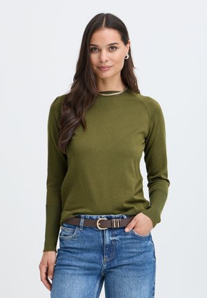 Femme aux longs cheveux foncés portant un pull vert olive et un jean bleu avec une ceinture marron, debout avec une main dans sa poche.