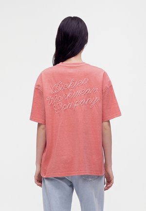 Person med langt mørkt hår iført en løs koralfarvet t-shirt med "Dickies Workwear Company" broderet på ryggen og lyseblå jeans.