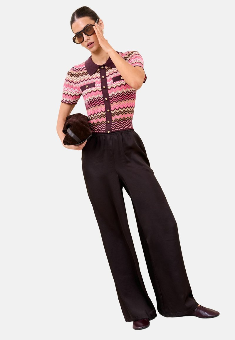 Femeie purtând ochelari de soare maro, bluză roz cu mâneci scurte și model, pantaloni largi negri, ținând o geantă plic maro în formă de pălărie.