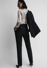 Pantaloni neri su misura abbinati a una blusa in seta beige chiaro, blazer nero appoggiato sulla spalla e scarpe con tacco alto nere.