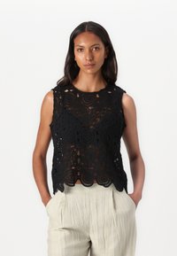 Svart heklet ermeløs topp med blomstermønstre og bølgekant. Kombinert med lys beige, plisserte bukser. Enfarget bakgrunn.