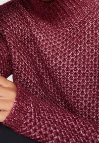 Maglione a maglia testurizzata color bordeaux con colletto alto e polsini a coste. Il materiale presenta un motivo intrecciato spesso, che offre calore e consistenza.