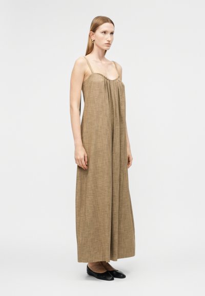 Γυναίκα στέκεται φορώντας ένα χαλαρό, αμάνικο, μακρύ φόρεμα σε ανοιχτό καφέ χρώμα με λεπτές τιράντες και μαύρα επίπεδα παπούτσια, μπροστά σε ένα απλό λευκό φόντο.