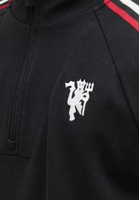 Μαύρο μπουφάν με φερμουάρ, με κεντημένο λευκό διάβολο που κρατά τρίαινα στο στήθος και κόκκινες και λευκές ρίγες στον ώμο.