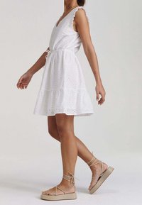 White cotton dress with eyelet detailing, V-neck, ruffled straps, and a tiered skirt. Paired with beige strappy sandals on a neutral background.