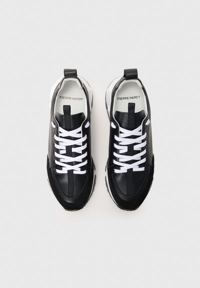 Zapatillas de cuero negro con cordones blancos, que presentan un diseño de panel texturizado, puntera redondeada y una lengüeta trasera prominente. Marca distintiva en el interior.