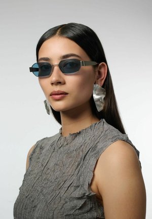 Femme aux longs cheveux bruns portant de petites lunettes de soleil rectangulaires bleues, de grandes boucles d'oreilles argentées abstraites et un haut gris texturé sans manches.