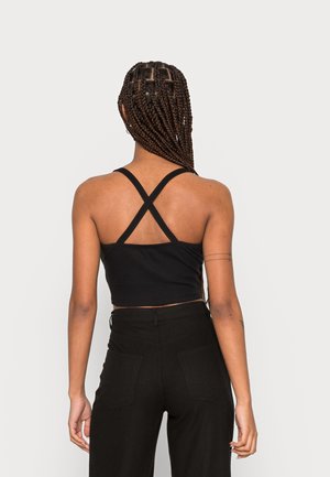 Schwarzes, kurz geschnittenes Tanktop mit überkreuzten Trägern. Das Material wirkt dehnbar und enganliegend. Kombiniert mit hoch taillierten schwarzen Hosen mit Gesäßtaschen.