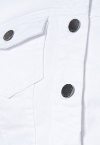 Primer plano de tela de mezclilla blanca con un bolsillo de solapa y tres botones metálicos plateados grabados con texto.