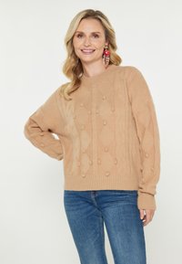 Uśmiechnięta kobieta z blond włosami nosi beżowy sweter z teksturowanej dzianiny, niebieskie dżinsy oraz duże czerwono-różowe kolczyki w kwiaty, pozując z ręką na biodrze.
