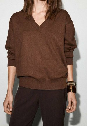 Vrouw draagt een bruine V-hals trui met opgerolde mouwen en donkere broek, geaccentueerd met twee grote bruine armbanden aan haar rechterpols.