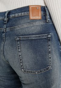 Tumma sininen farkut haalistuneella pinnalla, joissa on ruskea nahkapaikka vyötärölaudassa ja yksi takatasku. Klassinen viiden taskun muotoilu.