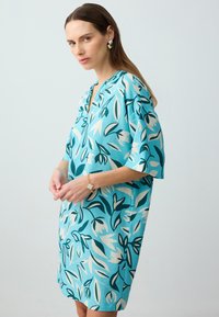 Un vestito morbido di color turchese con un motivo floreale, caratterizzato da maniche corte, scollatura a V e un design in tessuto testurizzato.