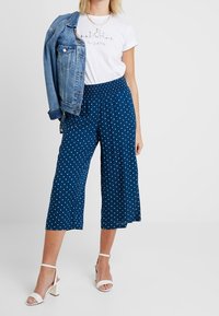 Granatowe spodnie culotte w biały groszek, elastyczny pas, połączone z białym t-shirtem z grafika i dżinsową kurtką, wykończone białymi sandałkami na obcasie.