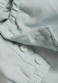 Svetlo modra tkanina z teksturirano površino, ki vsebuje bele gumbe in naborke ob robovih. Vidne podrobnosti šivov in šivanja.