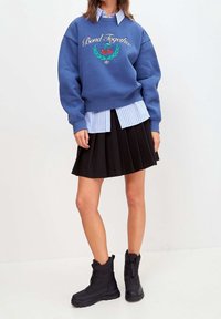 Sudadera azul con el estampado "Unidos por el vínculo" y un gráfico de cereza, superpuesta sobre una camisa de rayas azul y blanca, combinada con una falda negra con plisados y botas negras.