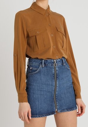 Donna che indossa una camicia marrone con bottoni e tasche sul petto, abbinata a una minigonna di jeans blu con cerniera frontale e anello di trazione.