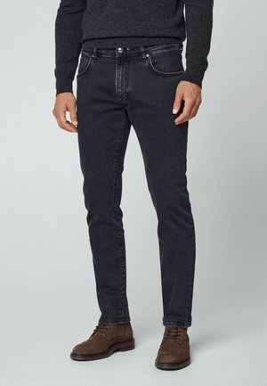 Mann in schmal geschnittener schwarzer Jeans, dunklem Pullover und braunen Wildleder-Schnürschuhen, der vor einem schlichten hellen Hintergrund steht.