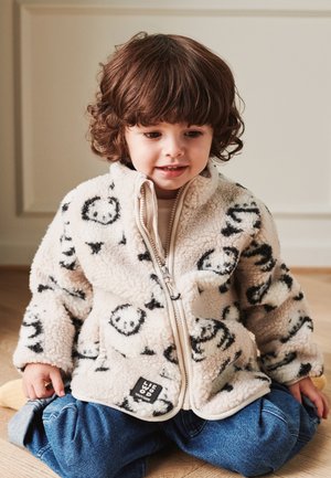 Karvane beež fleece-tagi mustade naeratavate mustritega, täislukuga, kõrge kraega ja valge äärisega. Kantud helepluusi ja siniste teksapükste peal.