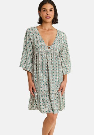 Femme portant une robe mi-longue avec des motifs ondulés verts et beiges, un décolleté en V profond et des manches trois-quarts, debout les mains détendues.
