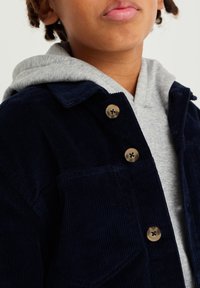 Námořnická kordurovová bunda s šedou mikinou pod ní. Čtyři knoflíkové zapínání a detail na hrudní kapse. Textura je žebrovaná a měkká.