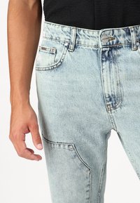 Jean en denim bleu clair avec une finition délavée, présentant un design à cinq poches et des accessoires en métal. Couture visible et coupe décontractée.