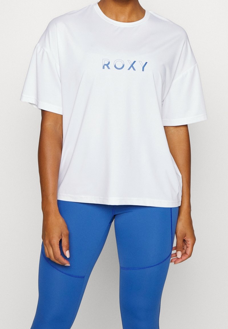 Camiseta blanca de manga corta con un logo azul de "ROXY", combinada con leggings azules. La tela es suave y ligeramente elástica.