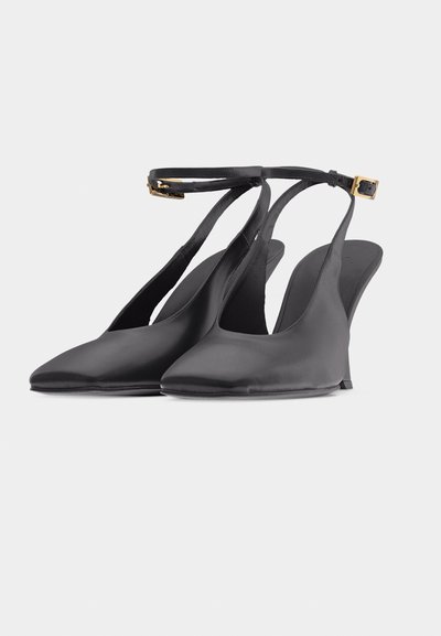 Μαύρα ψηλοτάκουνες γόβες με μυτερή μύτη, ανοιχτή πίσω πλευρά με λεπτά λουράκια στον αστράγαλο και χρυσές αγκράφες, σε λευκό φόντο.