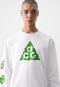 Long-sleeve white T-shirt featuring a green triangular leaf design with white lettering. Leaf motifs on sleeves add detail. Smooth fabric texture.