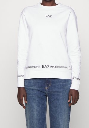 Osoba nosząca biały sweter z czarnymi logo Emporio Armani EA7 na klatce piersiowej, mankietach i dole, w połączeniu z niebieskimi dżinsami z denimu.