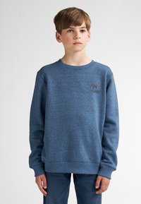 Sweat-shirt bleu en tissu polaire texturé, avec un col rond et des poignets côtelés. Petit logo sur la poitrine. Associé à un jean bleu.