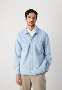 Light blue denim shirt with a button-up front, two chest pockets, and long sleeves. Features a classic collar and a relaxed fit.