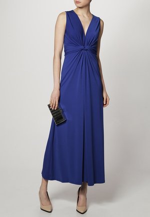 Frau trägt ein ärmelloses königsblaues Maxikleid mit einem verdrehten Knoten in der Taille, hält eine schwarze Clutch und trägt beigen Absatzschuhe.