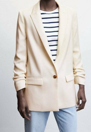 Blazer crème à un bouton, avec deux poches sur le devant et des manches retroussées, associé à une chemise rayée bleu marine et blanche, et un jean bleu clair.