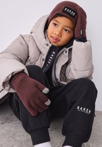 Dunjakke i lysegrå, sorte logodetaljer, brun strikbeanie og bordeaux handsker med grå spidser, båret med sorte joggebukser og hvide sokker.