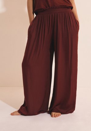 Femme portant un pantalon bordeaux, taille haute, jambes larges, avec une ceinture élastique et les mains dans les poches, debout pieds nus sur un sol clair.