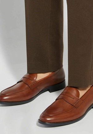 Mocassini in pelle marrone indossati con pantaloni marroni, caviglie visibili e suole nere su uno sfondo chiaro.