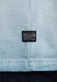 Tyrkysové bavlněné tričko s černým štítkem přišitým na spodní části, na kterém je bíle napsáno "Petrol Ind" ve stylu denimu.
