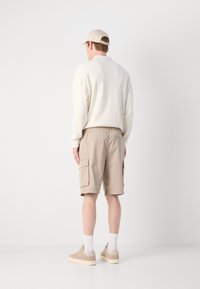 Bermudas bege com bolsos laterais, combinadas com um moletom creme e um boné bege. O conjunto inclui ténis bege e meias brancas.