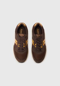 Baskets de sport marron avec des matériaux en maille et en cuir suédé, présentant des accents jaunes, des lacets plats et la marque "Saucony" imprimée sur la languette.