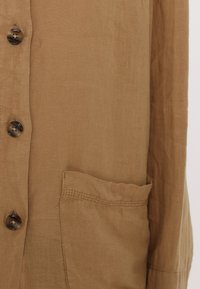 Brown linen shirt featuring a button-down front, large buttons, and a side pocket with stitched detailing. Soft, textured fabric.