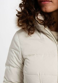 Giaccone imbottito beige chiaro con colletto alto, chiusura con zip e texture trapuntata. Tessuto morbido con una finitura liscia e leggermente lucida.