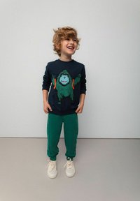 Granatowa bluza z grafiką zielonej goryla, zestawiona z zielonymi dresami i białymi trampkami z beżowymi sznurówkami. Gładkie, casualowe ubranie.