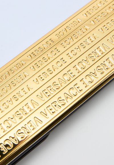 Zelta metāla aksesuārs ar izspiedumu "VERSACE" tekstu, gluda tekstūra, taisnstūrveida forma un spīdīgs, tīru malu apdare.