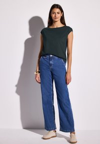 Vihreä lyhythihainen paita yhdistettynä korkeavyötäröisiin sinisiin farkkuleggingseihin. Vaaleanruskeat sneakersit, joissa on teksturoitu pinta. Yksinkertainen muotoilu.