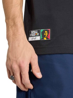 Κοντινό πλάνο ενός χεριού που φοράει μαύρο δαχτυλίδι, δίπλα σε ένα μαύρο πουκάμισο με ένα αυτοκόλλητο που γράφει «Το ποδόσφαιρο είναι ελευθερία» και ένα πολύχρωμο πορτρέτο.