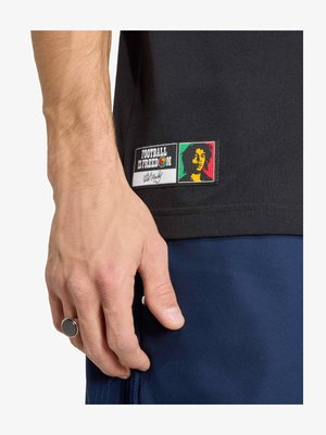 Κοντινό πλάνο ενός χεριού που φοράει μαύρο δαχτυλίδι, δίπλα σε ένα μαύρο πουκάμισο με ένα αυτοκόλλητο που γράφει «Το ποδόσφαιρο είναι ελευθερία» και ένα πολύχρωμο πορτρέτο.